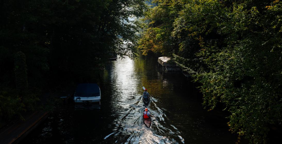 Two people paddle in canoes in a narrow river. 