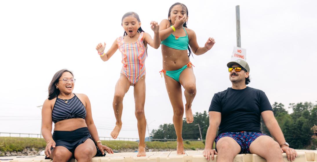 Two young girls jump off a dock while their parents watch. 