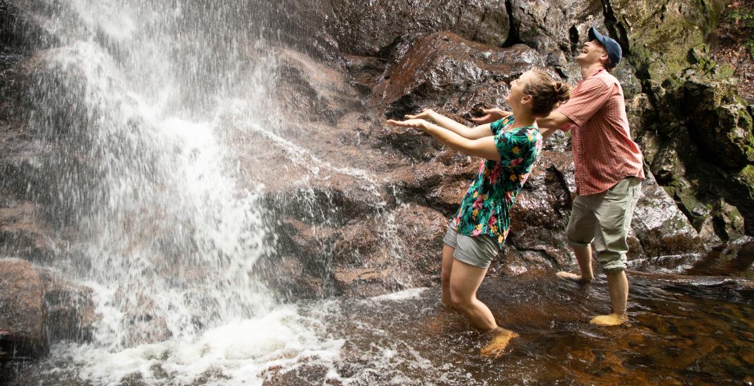Two young people stand beneath a waterfall.