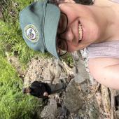 A woman wearing a baseball cap smiles in front of a rocky trail.