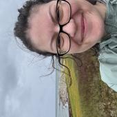 A woman with windblown hair smiles in front of a grey coastline.