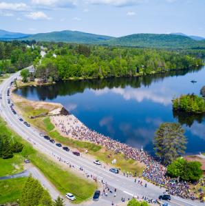 Aerial view of a lake bordered by lush green forest and a winding road. A large group of cyclists gathers near the road