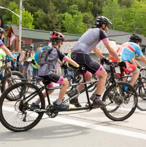 Cyclists race on a sunlit gravel road surrounded by lush green trees. The lead riders appear focused and determined