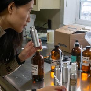 A woman smells a small silver bottle while surrounded by various amber-colored bottles and other containers of liquid on a stainless steel counter.