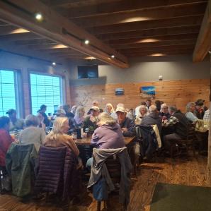 A large group of people are seated at long tables in a restaurant with wood walls and a high ceiling.