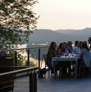 A large group of people sit at a long table set for dining on a deck overlooking a body of water with the Adirondack mountains in the background.