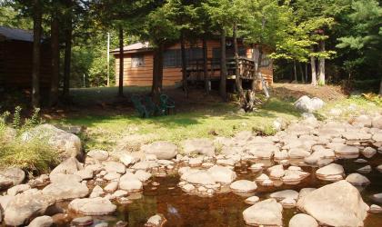 An image of a quaint cottage surrounded with trees and a rocky rivers edge.