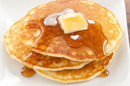 a stack of three pancakes on a white plate with a tab of butter on top and syrup dripping down the sides