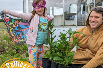 A little girl with butterfly wings on and a butterfly mask standing next to a mand holdong potted plants that are good for pollinators