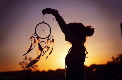 A silhouette of a woman holding up a dream catcher with the sunsetting in the background 