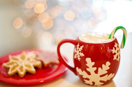 A photo with a couple snowflake cookies on a red plate next to a red mug with white snowflakes with hot cocoa and a candycane in it