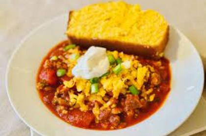 A bowl of chili with shredded cheddar cheese and sour cream on top with a side of corn bread