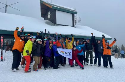 A large group of skiers and snowboarders in winter gear cheer and wave while holding an I Love NY LGBTQ+ pride flag.