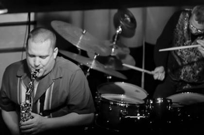 A photo of Keith plaung the saxophone with some else in the background playing on a drum set