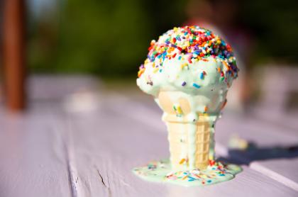 Ice cream cone with green ice cream and rainbow sprinkles standing on a picnic table with melting drips running down the side