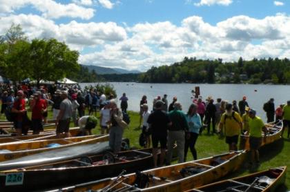 Large group of people wait on the green by the water with their canoes