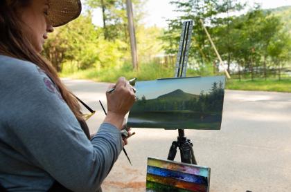 A woman in a blue long sleeve shirt and sun hat is out side painting the landscape that is before her