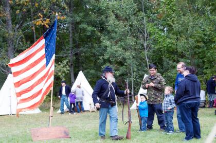 A man in a Union soldier uniform carrying a musket and talking with people attending the event. An American flag of that era is flying on a pole and there are white canvas tents in the background