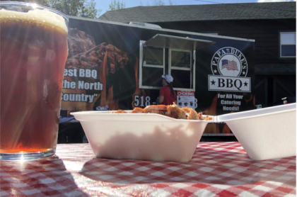 A tabletop photo of a full glass of beer and an open container of BBQ food on a picnic table woth red and white gingham table cover