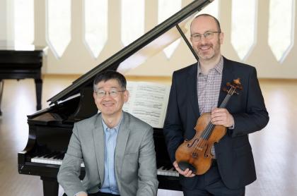 A man sitting on the bench in front of a grand piano with a man standing next to him holfing a violin