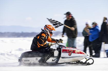 A snowmobiler riding past a man waving a checkered flag at the end of the race