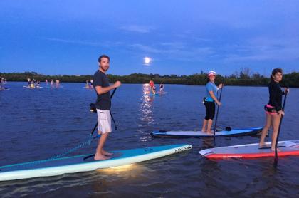 Three people on stand-up paddle boards paddleing by moon light