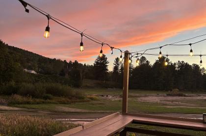 view from a wooden deck with party lights strung above looking out to trees and a sunset with orange and pink colored clouds