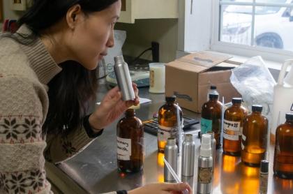 A woman smells a small silver bottle while surrounded by various amber-colored bottles and other containers of liquid on a stainless steel counter.