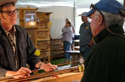 A man in a dark jacket and hat shows a handcrafted fishing rod to two people.
