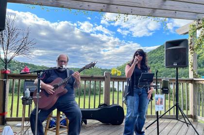 A man and woman perform music on an outdoor deck. The man plays an acoustic guitar and the woman sings into a microphone. They are positioned in front of a scenic view and audio equipment.