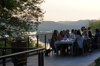 A large group of people sit at a long table set for dining on a deck overlooking a body of water with the Adirondack mountains in the background.