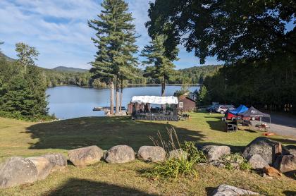 Outdoor stage set up on a lakeside lawn with a few cars parked nearby.