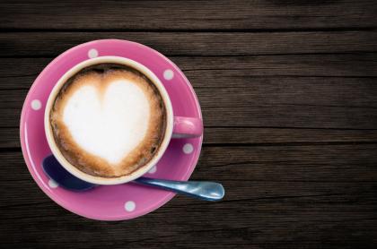 A cappuccino with heart-shaped foam sits in a pink polka-dot cup and saucer on a wooden table
