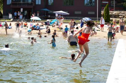 A girl in a red swimsuit jumps into a lake at a crowded beach.  Many people are swimming