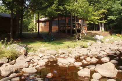 An image of a quaint cottage surrounded with trees and a rocky rivers edge.