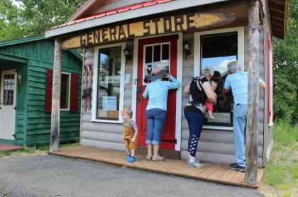 Adults and small children on the porch of a miniature wooden general store.