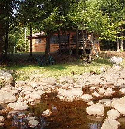 An image of a quaint cottage surrounded with trees and a rocky rivers edge.