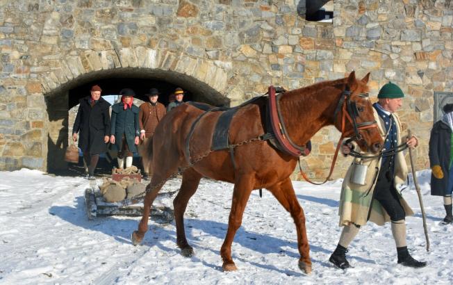 People in colonial period clothes lead a horse pulling a sled in a stone fort.
