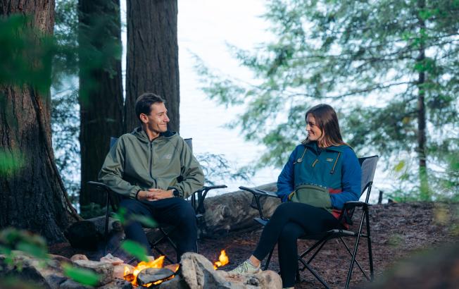 A man and woman sit in camp chairs at a campfire.