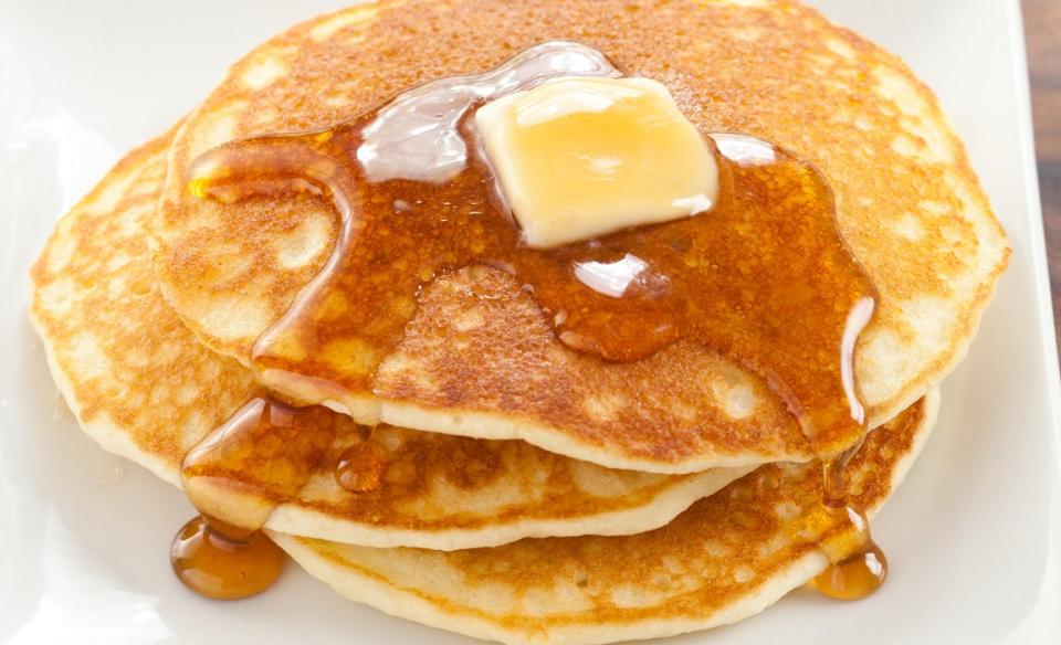 a stack of three pancakes on a white plate with a tab of butter on top and syrup dripping down the sides