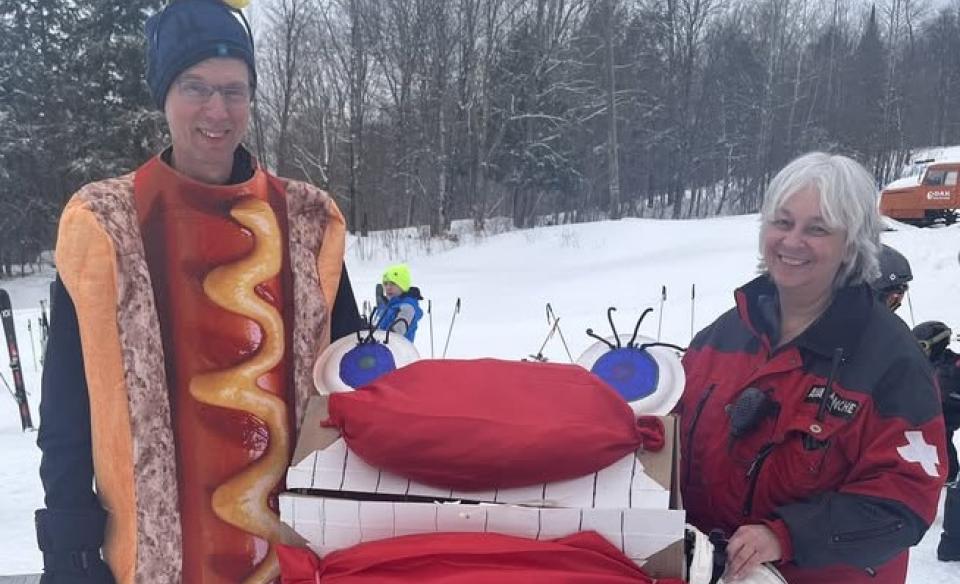 A mad dressed up on a hot dog costume as he and a women hold up a silly cardboard sled