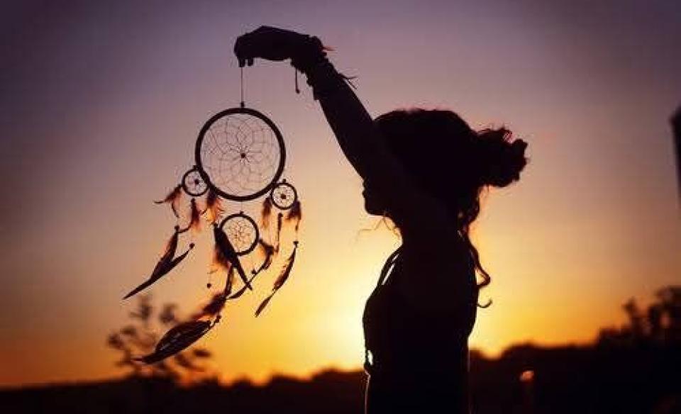A silhouette of a woman holding up a dream catcher with the sunsetting in the background
