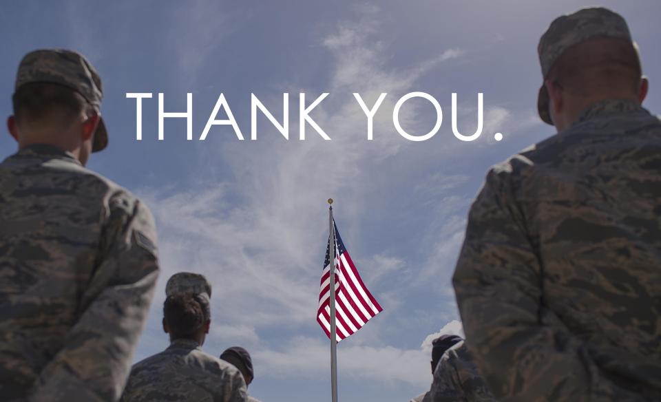 A photo of several military members standing facing an American flag in front of them with the words Thank You. typed in the sky