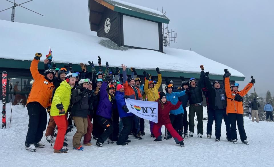 A large group of skiers and snowboarders in winter gear cheer and wave while holding an I Love NY LGBTQ+ pride flag.