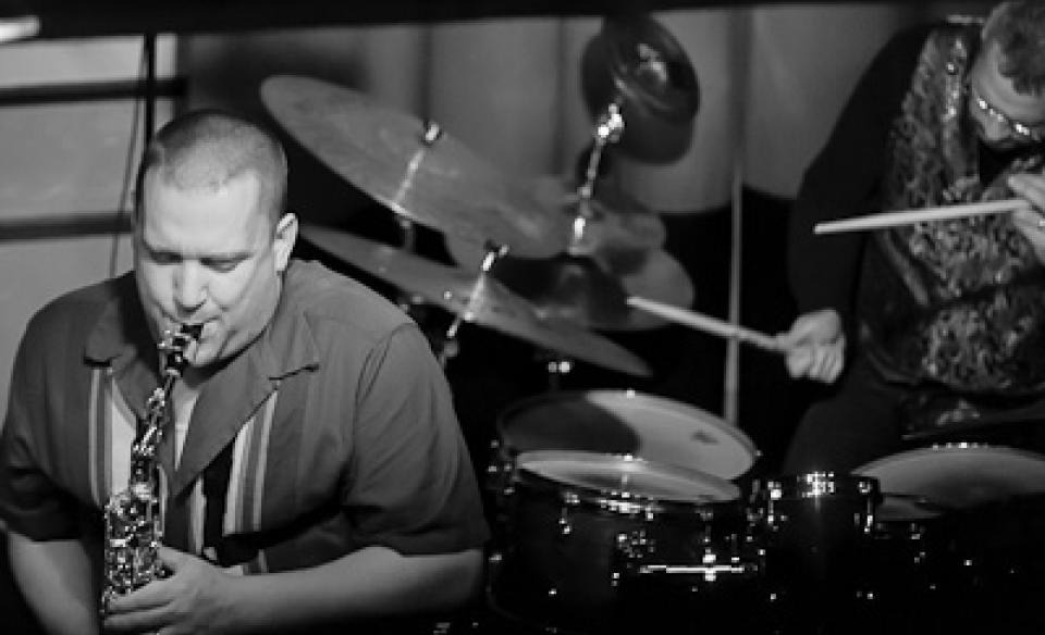 A photo of Keith plaung the saxophone with some else in the background playing on a drum set