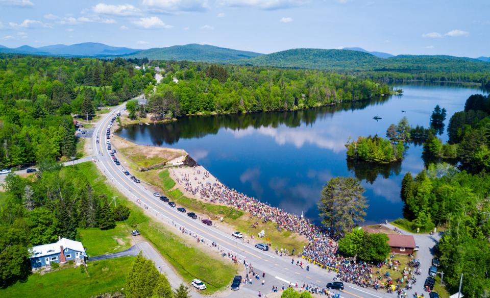 Aerial view of a lake bordered by lush green forest and a winding road. A large group of cyclists gathers near the road