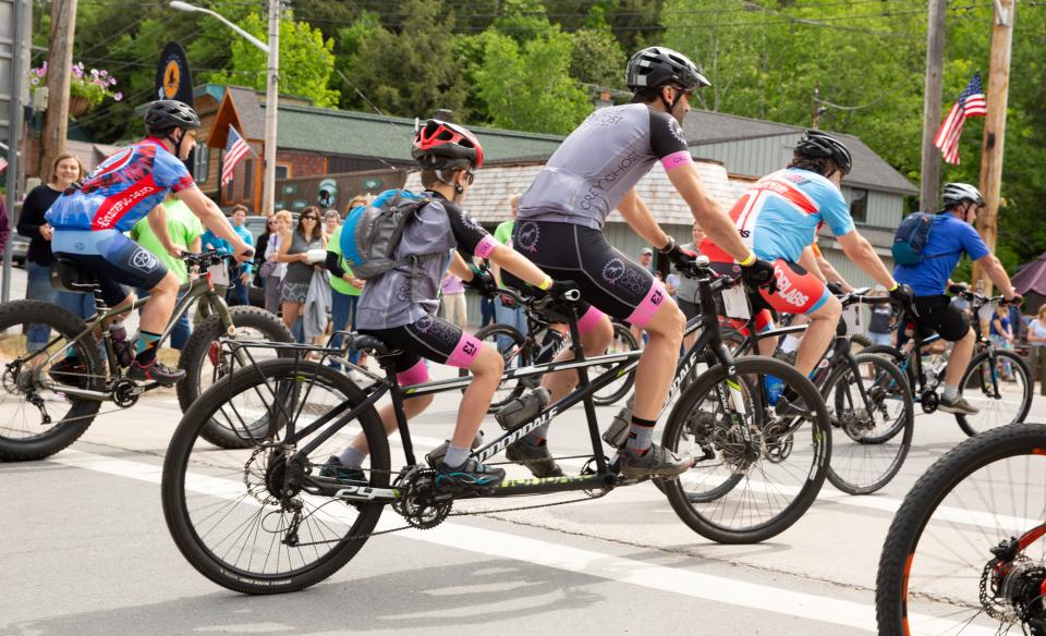 Cyclists race on a sunlit gravel road surrounded by lush green trees. The lead riders appear focused and determined