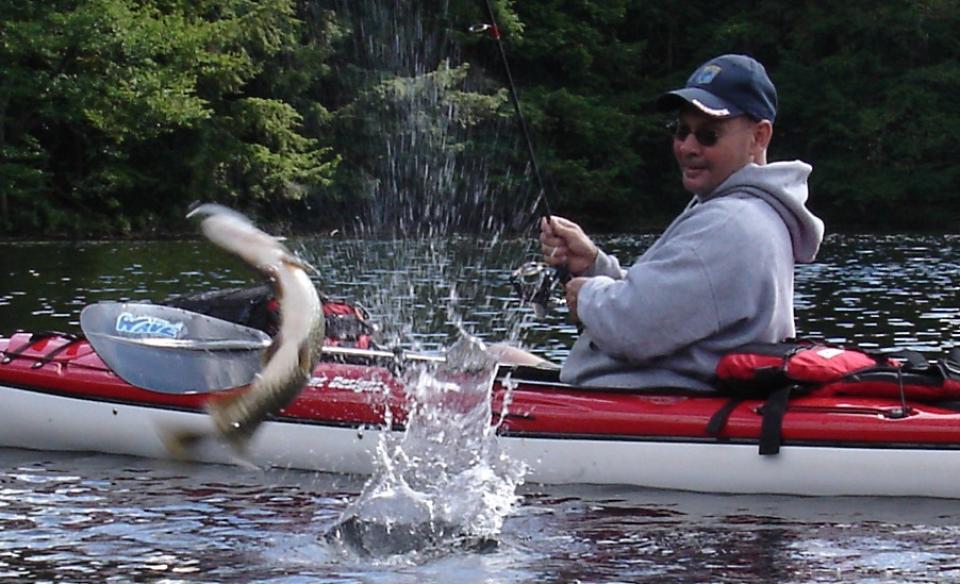 Man in kayak fishing for pike as the fish jumps out of the water on his line.