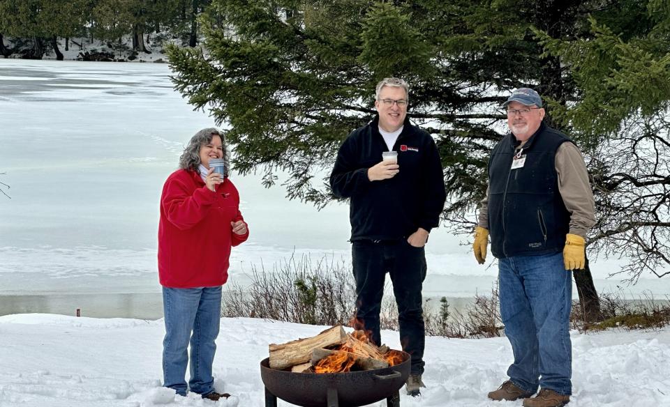 Three people standing around a campfire outside in the snow drinking warm beverages
