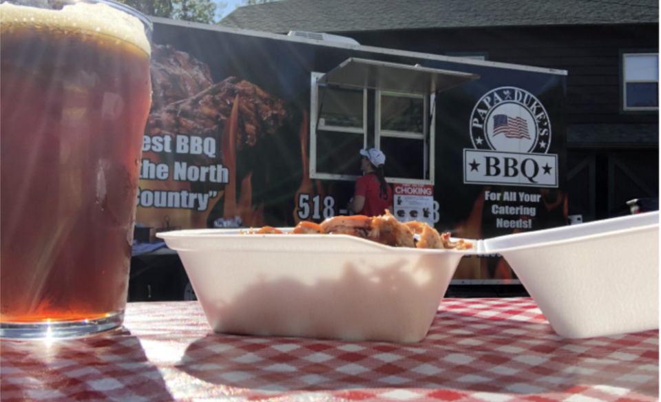A tabletop photo of a full glass of beer and an open container of BBQ food on a picnic table woth red and white gingham table cover